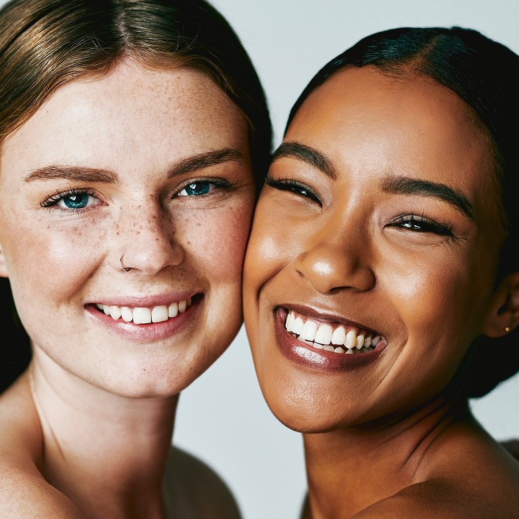 Two teenage girls, one light-skinned with freckles and red hair, the other dark-skinned with dark hair, smile with their cheeks touching, conveying a sense of youthful vibrancy and friendship.